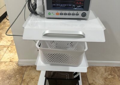 A medical EKG monitor on a wheeled cart with visible cables and a plastic basket beneath it, placed on a tiled floor.