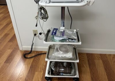 A medical EKG monitor on a wheeled cart with visible cables and a plastic basket beneath it, placed on a tiled floor.